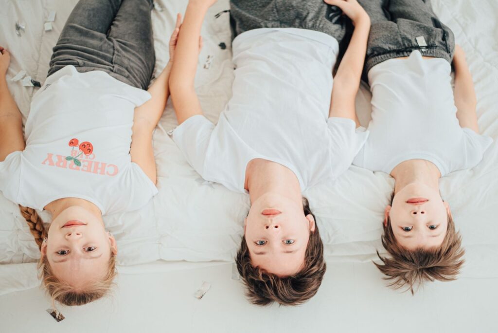 Children lying on a white bed, wearing casual white t-shirts. Bright and playful indoor setting.