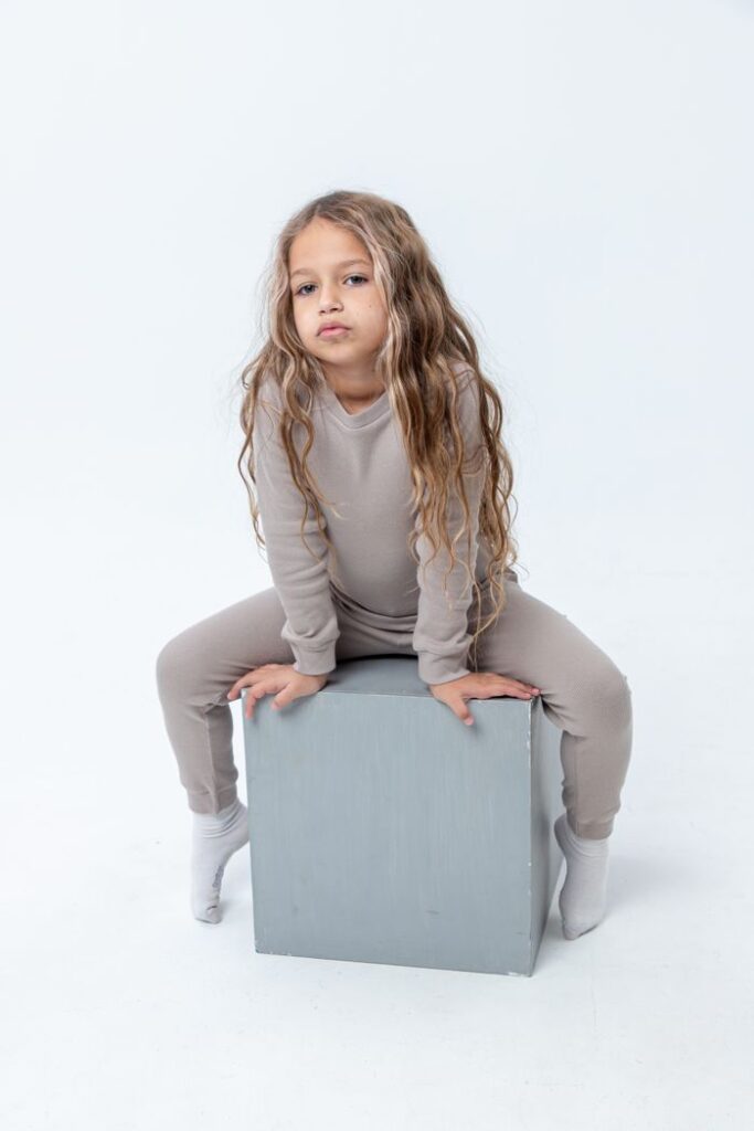 Child with long blonde hair sitting on a box in a studio with a white background.