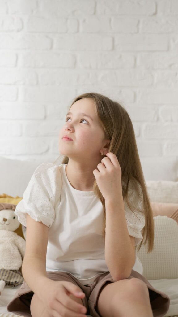 A pensive girl sits indoors, looking upwards, surrounded by soft toys. Captures childhood innocence.